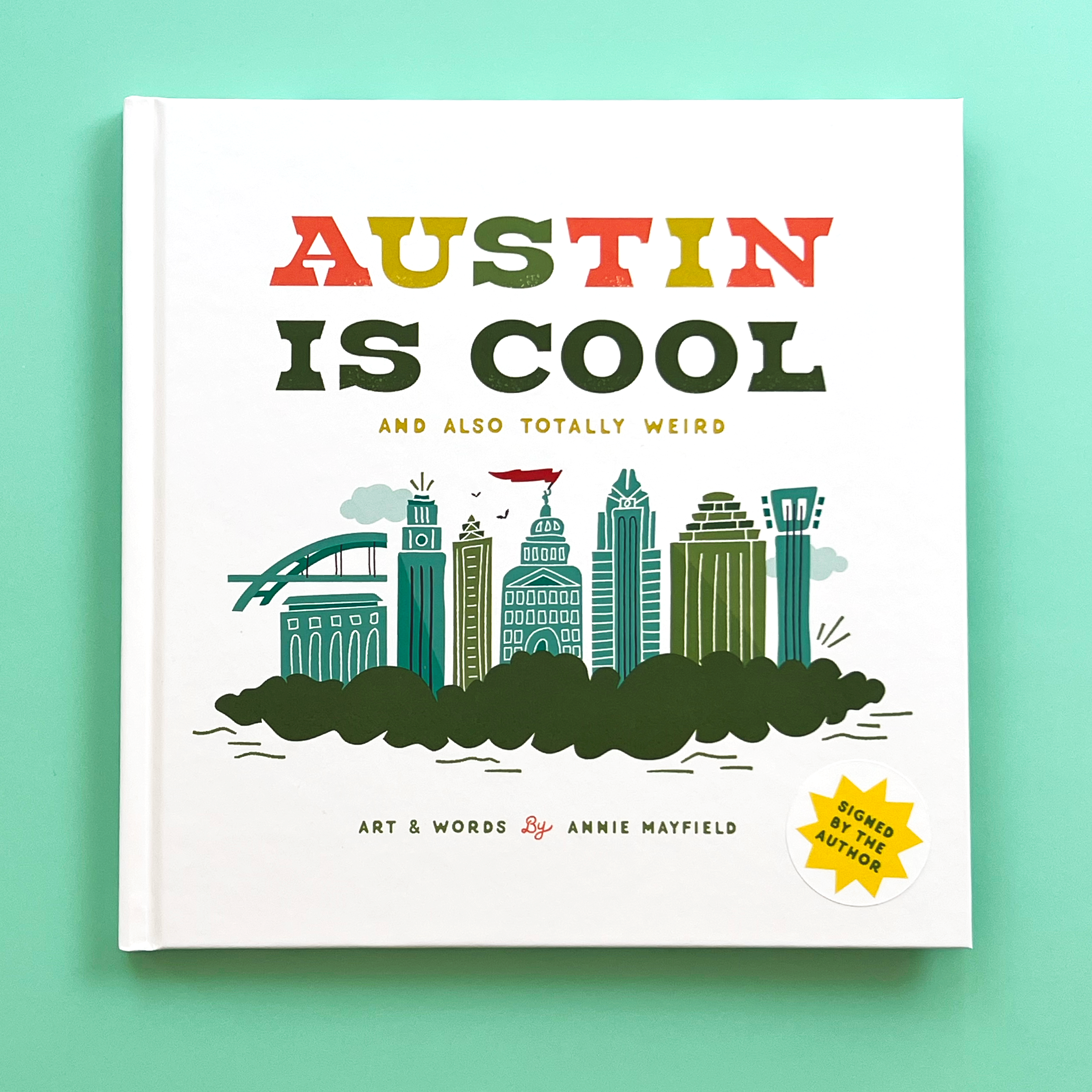 A book titled 'Austin Is Cool' featuring a colorful illustration of the Austin cityscape with various iconic landmarks, written by Austinite, Annie Mayfield.